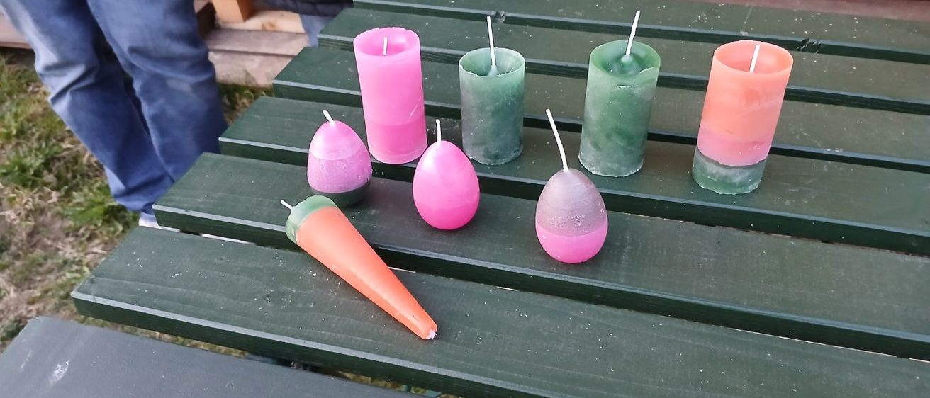 Auf einem Picknicktisch steht eine Reihe von Kerzen in verschiedenen Farben und Formen, darunter rosa, grün und orange. Eine Person steht hinter dem Tisch.