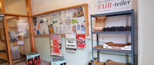 Ein Raum mit einem Bulletin-Board, einem Regal mit Aufbewahrungsboxen und einem Schild für 'Fair-Teiler'. Zwei Boxen mit 'Plat da'-Etiketten liegen auf dem Boden.
