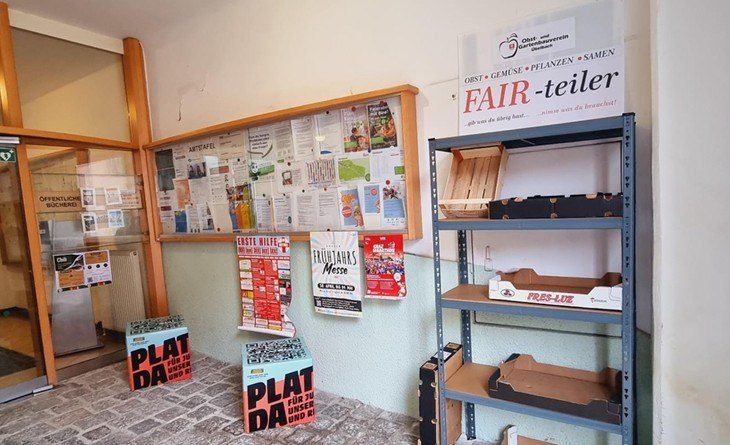 Ein Raum mit einem Bulletin-Board, einem Regal mit Aufbewahrungsboxen und einem Schild für 'Fair-Teiler'. Zwei Boxen mit 'Plat da'-Etiketten liegen auf dem Boden.