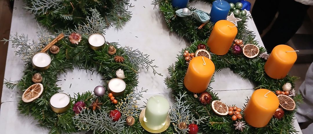 Ein Tisch zeigt verschiedene Kränze, geschmückt mit immergrünen Pflanzen, Kerzen in verschiedenen Farben sowie saisonalen Früchten und Nüssen. Ein Kranz hat ein lilafarbenes Band.