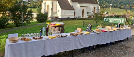 Vor einer Kirche mit einem grünen Rasen und einem Baumstumpf ist ein Buffettisch mit verschiedenen Speisen und Getränken aufgebaut.