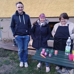 Drei Menschen stehen vor einem Haus mit Kerzen auf einem Tisch. Der Mann trägt eine Kapuze, die Frauen tragen Wintermäntel und Schürzen. Dahinter steht eine Bank.