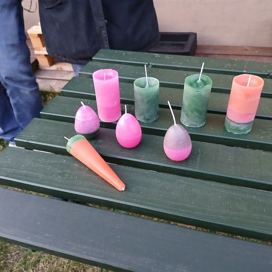 Ein grüner Tisch hat sieben Kerzen. Drei sind rosa, drei sind grün und eine ist orange. Eine Person in Jeans steht hinter dem Tisch.