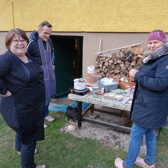 Drei Personen stehen draußen in der Nähe eines Gebäudes, mit einem Tisch, auf dem Kochutensilien stehen, und einem Holzhaufen dahinter. Die Frau rechts trägt eine Bandana und einen Pelzmantel.