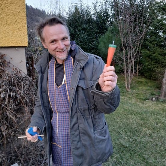 Ein Mann in einer grauen Jacke hält draußen an einem sonnigen Tag eine Karottenkerze. Er lächelt und hält die Karotte in seiner rechten Hand.