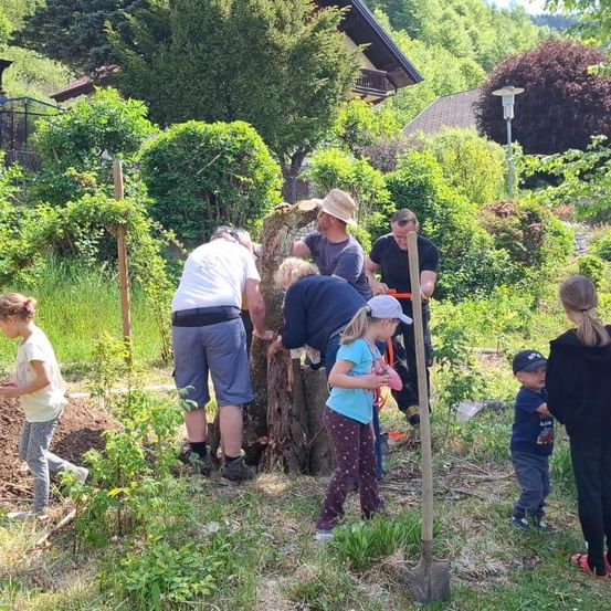 Eine Gruppe von Menschen pflanzt einen Baum in einem Garten. Einige tragen Hüte und Schuhe. Ein kleines Mädchen hält eine Schaufel. Ein Mann hält einen Baumstumpf. Ein Junge und eine Frau stehen neben dem Mann.