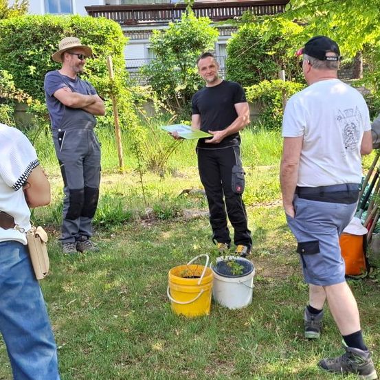 Eine Gruppe von Menschen steht in einem Garten, zwei von ihnen halten ein Stück Papier. Ein Mann trägt einen Hut und der andere einen Rucksack.