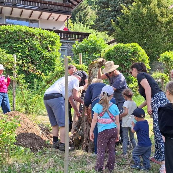 Eine Gruppe von Erwachsenen und Kindern arbeitet gemeinsam in einem Garten, wobei einige Menschen graben und andere beobachten. Ein Haus ist im Hintergrund zu sehen.