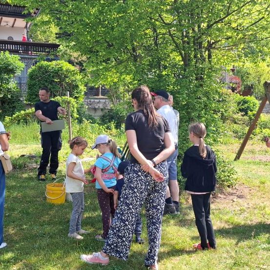 Eine Gruppe von Menschen steht in einem Garten, ein Mann hält ein Klemmbrett, Kinder halten Eimer und eine andere Frau beobachtet. Dahinter sind ein Haus und Bäume zu sehen.