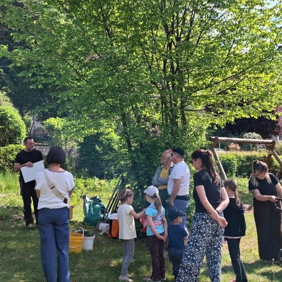 Eine Gruppe von Menschen versammelt sich in einem Garten, ein Mann hält ein Papier und spricht. Ein paar Kinder stehen in der Nähe, und einige Erwachsene stehen hinter ihnen. Es gibt Eimer, Taschen und Stöcke auf dem Boden.