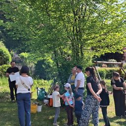 Eine Gruppe von Menschen versammelt sich in einem Garten, ein Mann hält ein Papier und spricht. Ein paar Kinder stehen in der Nähe, und einige Erwachsene stehen hinter ihnen. Es gibt Eimer, Taschen und Stöcke auf dem Boden.