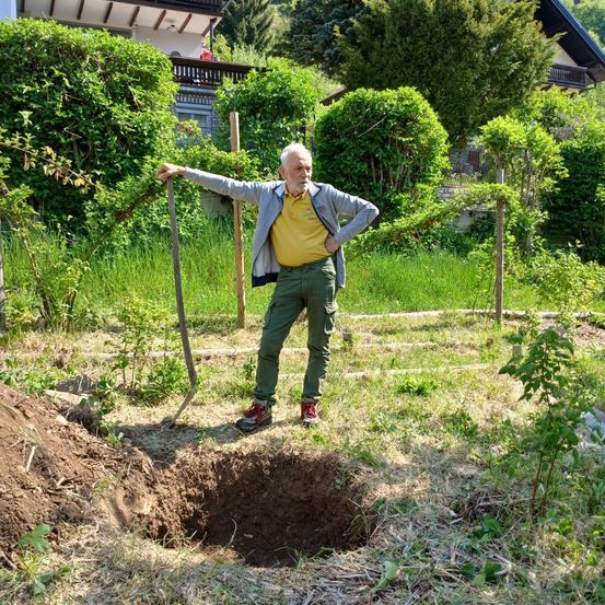 Ein älterer Mann in einem gelben Shirt steht in einem Garten mit einer Schaufel neben einem Loch. Dahinter sind Büsche, ein Haus und ein Balkon.