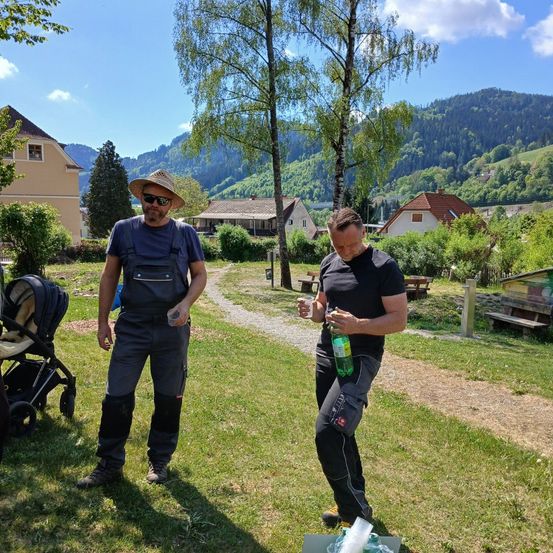Zwei Männer in Freizeitkleidung, einer mit Hut, stehen auf einem Rasen. Einer hält eine grüne Flasche und der andere schaut nach unten. Ein Kinderwagen und Bänke sind im Hintergrund.