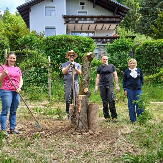 Vier Personen stehen in einem Garten vor einem Haus, halten Werkzeuge und lächeln. Eine Person hält eine Harke, eine andere einen abgesägten Baumstumpf.
