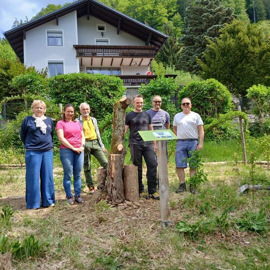 Fünf Personen stehen vor einem Haus mit einem geschnittenen Baumstumpf. Ein Mann hält ein Schild.