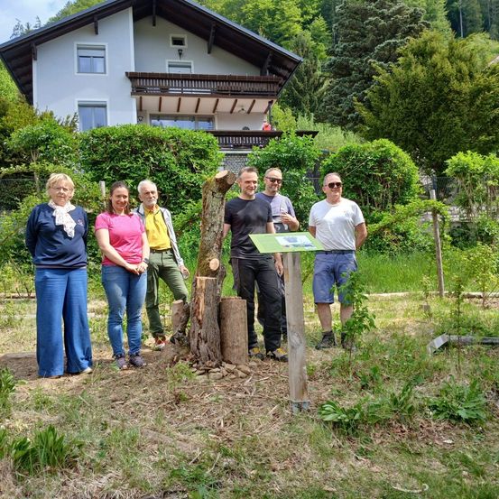 Fünf Personen stehen vor einem Baumstumpf in einem Garten mit einem großen Haus im Hintergrund.