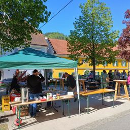 Ein Markt ist unter einem grünen Zelt mit verschiedenen Artikeln auf Tischen aufgebaut. Leute gehen herum und einige sitzen an Tischen. Gebäude sind im Hintergrund.