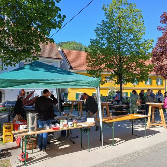 Ein Markt ist unter einem grünen Zelt mit verschiedenen Artikeln auf Tischen aufgebaut. Leute gehen herum und einige sitzen an Tischen. Gebäude sind im Hintergrund.
