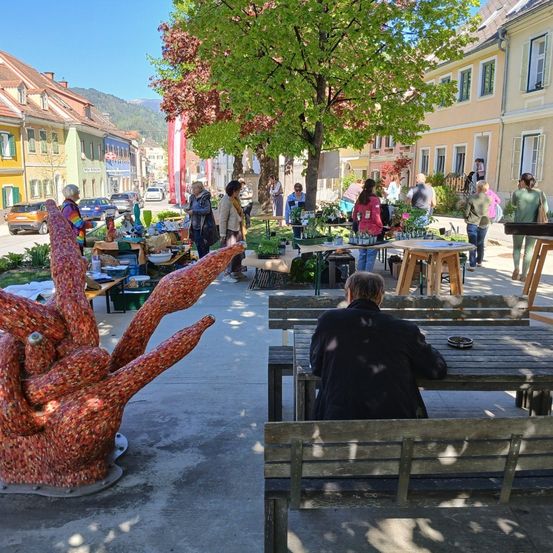 Ein geschäftiger Markt ist auf einem Stadtplatz aufgebaut, mit Leuten, die verschiedene Stände unter einem großen Baum erkunden. Bunte Kunstwerke sind ausgestellt und ein Mann sitzt auf einer Bank. Gebäude säumen die Straße im Hintergrund.
