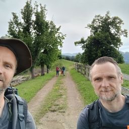 Zwei Männer mit Rucksäcken posieren auf einem ländlichen Pfad mit Menschen im Hintergrund. Bäume und ein Holzzaun säumen den Weg.