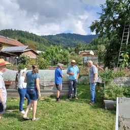 Eine Gruppe von Menschen in einem Garten, einige tragen Hüte, mit einem bergigen Hintergrund. Sie stehen im Kreis und diskutieren möglicherweise etwas.