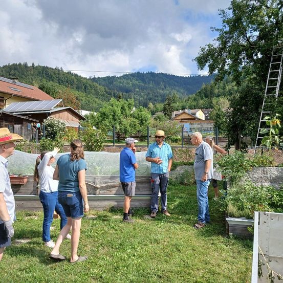 Eine Gruppe von Menschen in einem Garten, einige tragen Hüte, mit einem bergigen Hintergrund. Sie stehen im Kreis und diskutieren möglicherweise etwas.