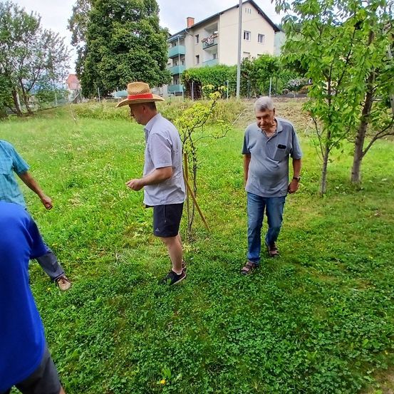 Drei Männer gehen durch ein saftiges Feld mit einem kleinen Baum im Vordergrund. Ein Mann trägt einen Strohhut und ein weißes Hemd. Ein anderer Mann trägt ein graues Hemd und Jeans. Ein dritter Mann trägt ein blaues Hemd.
