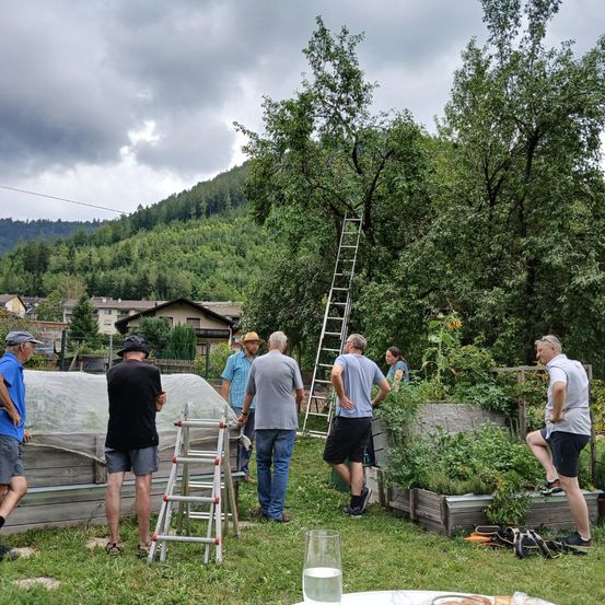 Eine Gruppe von Menschen in einem Garten mit einem Berg im Hintergrund, ein Mann lehnt sich an eine Leiter, während andere stehen und reden.