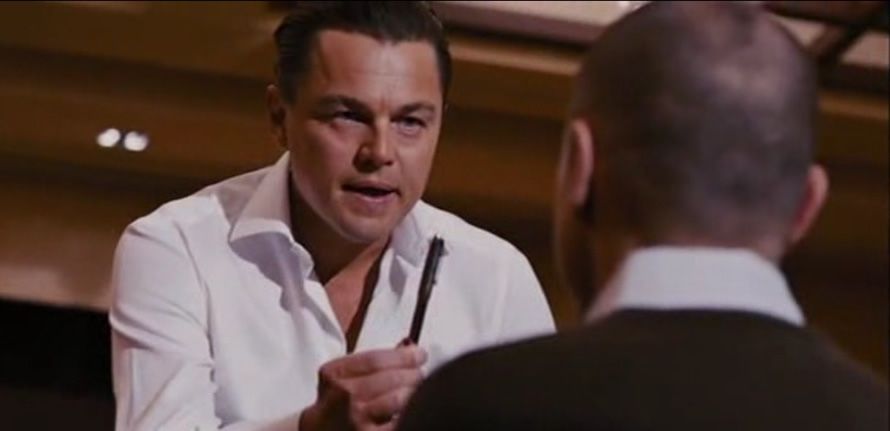 Leonardo DiCaprio, eine Feder in der Hand, scheint sich in einem ernsthaften Gespräch mit einem anderen Mann in einem schwarzen Anzug zu befinden.