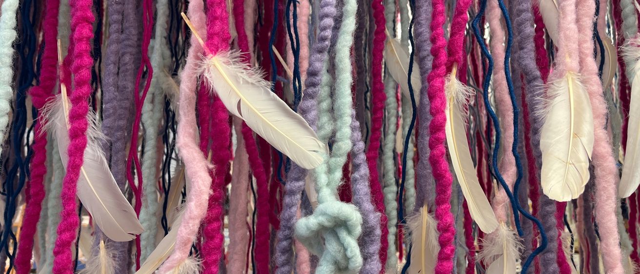 Eine farbenfrohe Ausstellung von Garn und Federn, mit rosa, lila, blauen und weißen Fäden und Federn, die in einem Cluster hängen.