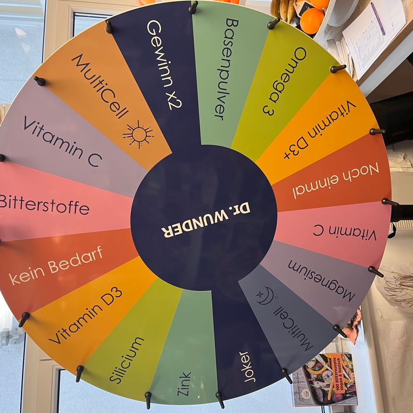 Ein Drehrad zeigt verschiedene ernährungsbezogene Kategorien, einschließlich Vitamin C, Vitamin D3, Magnesium, Omega 3 und mehr. Die Mitte ist mit 'MEGANE 13' markiert. Das Rad ist von zusätzlichen lebensmittelbezogenen Artikeln umgeben.