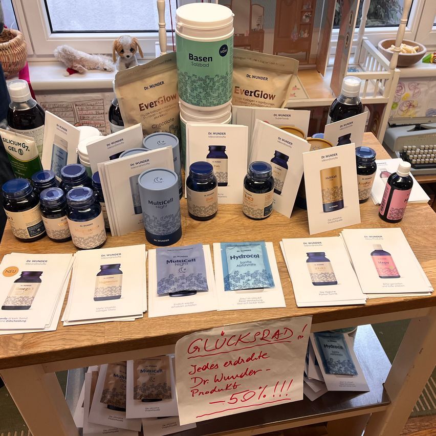 Ein Holztisch mit verschiedenen Flaschen und Produkten, darunter 'Basen' und 'EverGlow'. Es gibt Werbematerialien, ein Schild mit 'Glucksrad' und Artikel wie MultiCell Night. Ein Kuscheltier und Spielzeug im Hintergrund.