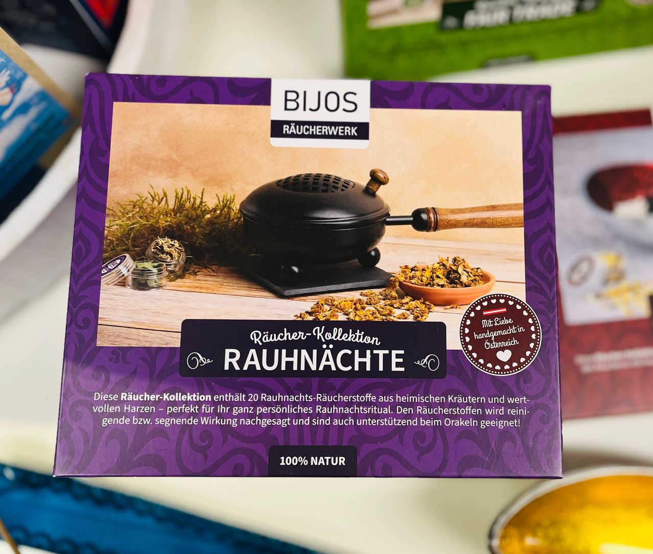 Eine lila Box der Bijos Räucherwerk-Kollektion. Die Vorderseite zeigt ein Bild eines Räucherbrenners mit Kräutern und Blumen, beschriftet mit 'Räucher-Kollektion Räuchnachte' und einem Herzsymbol. Das Produkt ist als 100% natürlich zertifiziert.