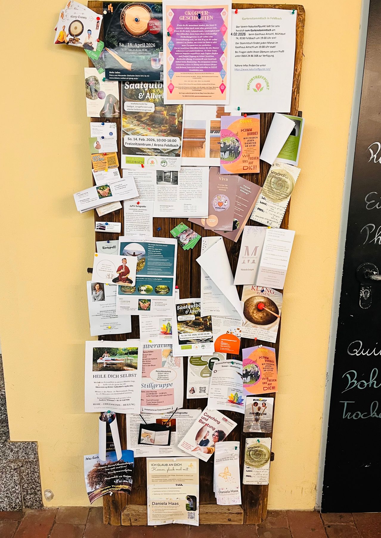 Eine Pinnwand zeigt verschiedene Werbungen, Flugblätter und Mitteilungen. Sie beinhaltet ein Flugblatt mit dem Titel 'Saatgut & Alter' und ein weiteres mit einem Bild einer Person und den Worten 'HEIL DIECH SELBST'.