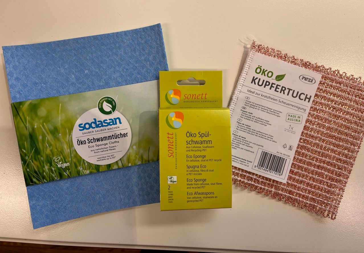 Three eco-friendly cleaning products on a white surface: a blue sponge cloth, a yellow box of sponges, and a red and white cloth.