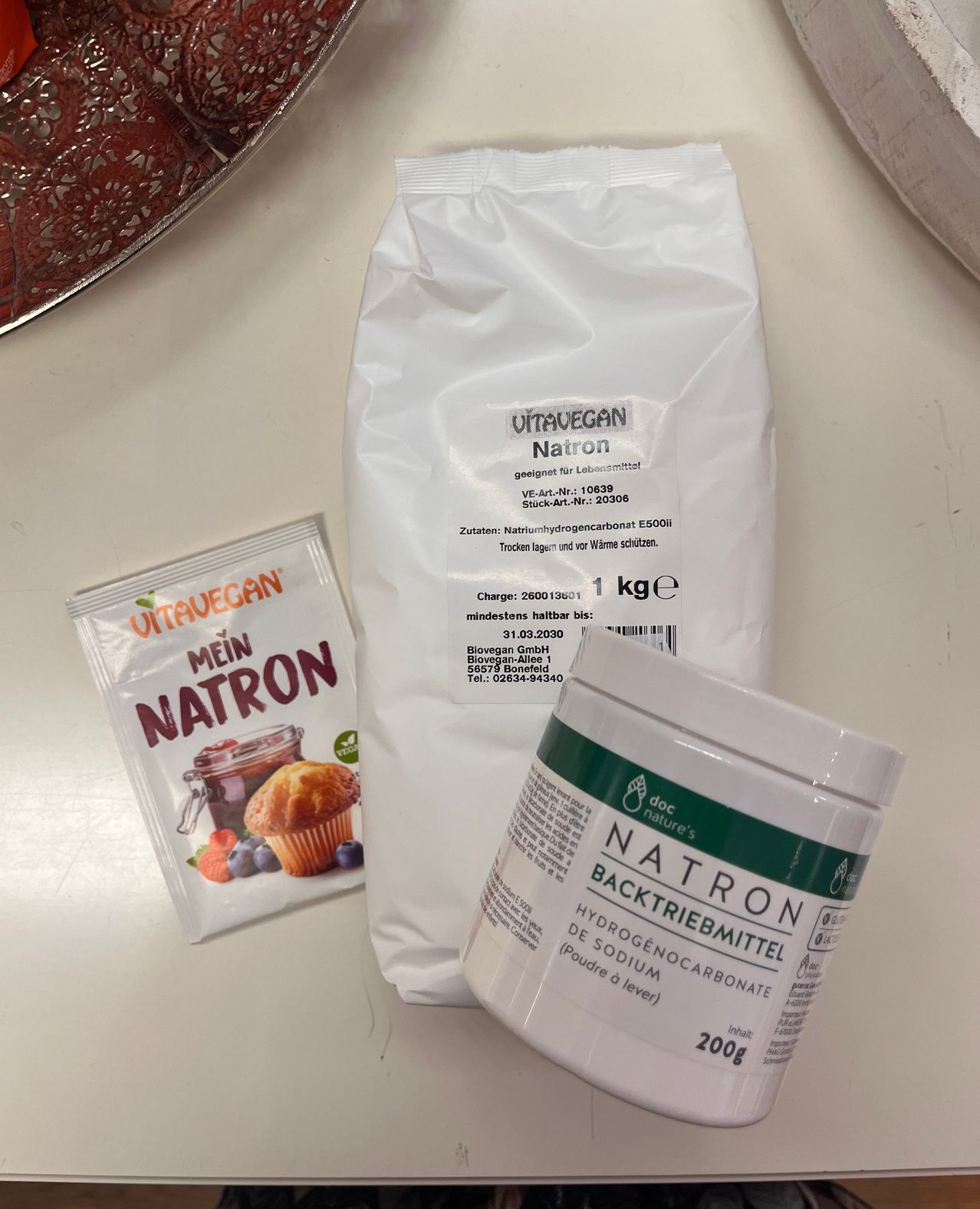 A white bag of baking soda, a small packet of baking soda, and a container of baking soda are on a table.
