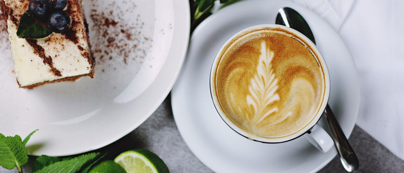 Ein weißer Teller mit einem Stück Kuchen und eine Tasse Kaffee mit einem Blattdesign auf einer Untertasse. Limetten und Minzblätter sind auf dem Tisch.