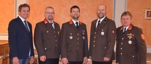 Fünf Männer stehen lächelnd für ein Foto in formeller Kleidung. Sie tragen braune Uniformen mit roten Säumen und Medaillen. Ein orangefarbener Hintergrund und ein Holzboden sind zu sehen.