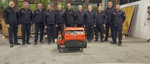 Eine Gruppe von Feuerwehrleuten posiert für ein Foto mit einer orangefarbenen Maschine in der Mitte, alle tragen schwarze Uniformen.