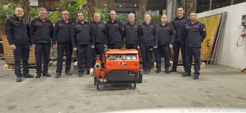 Eine Gruppe von Feuerwehrleuten posiert mit einer orangefarbenen Maschine in einem Lager, umgeben von Topfpflanzen und einem gelben Banner.