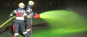 Zwei Feuerwehrleute in Schutzkleidung spritzen grüne Flüssigkeit aus einem Feuerlöscher auf eine Rasenfläche.