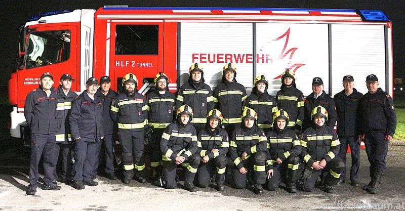 Eine Gruppe von Feuerwehrleuten in Uniform posiert für ein Foto vor einem Feuerwehrwagen. Sie tragen alle Helme, Handschuhe und Stiefel.