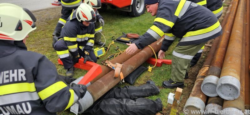Eine Gruppe von Feuerwehrleuten in Uniform und Helmen arbeitet an einer großen Rohrleitung auf einem Rasenfeld. Ein Feuerwehrmann beugt sich über die Rohrleitung, während andere ihm helfen.