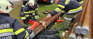 Eine Gruppe von Feuerwehrleuten in Uniform und Helmen arbeitet an einer großen Rohrleitung auf einem Rasenfeld. Ein Feuerwehrmann beugt sich über die Rohrleitung, während andere ihm helfen.