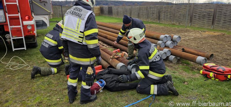 Eine Gruppe Feuerwehrleute in Uniform arbeitet an einer großen Rohrleitung auf einem Grasfeld, während ein anderer Feuerwehrmann auf dem Boden liegt.