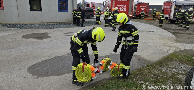 Feuerwehrleute versammeln sich vor einem Gebäude, einer bereitet einen Schlauch vor. Andere stehen mit Feuerwehrwagen im Hintergrund. Die Szene spielt sich auf einem gepflasterten Bereich mit einem Grasfleck ab.