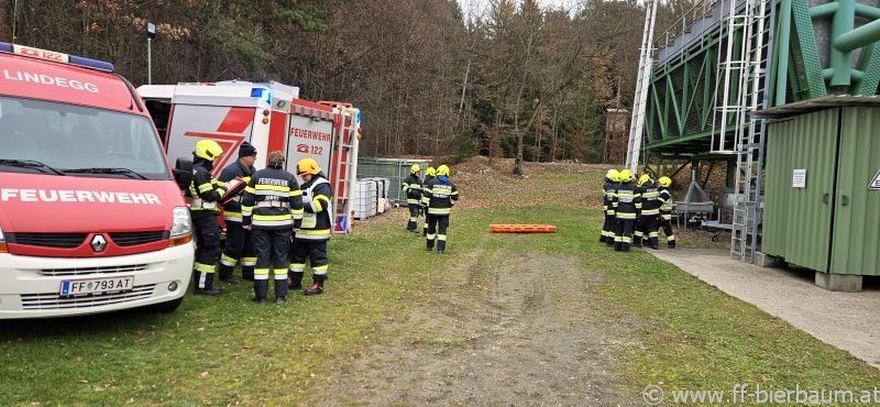 Eine Gruppe von Feuerwehrleuten versammelt sich auf einem Feld, mit Feuerwehrfahrzeugen und Ausrüstung sichtbar. Sie tragen gelbe Helme und schwarze Uniformen.