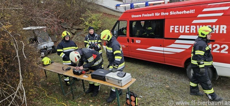 Eine Gruppe von Feuerwehrleuten in voller Ausrüstung arbeitet an Geräten an einem Tisch. Ein roter Feuerwehrwagen ist hinter ihnen geparkt. Das Gebiet ist von Pflanzen und einem Golfwagen umgeben.