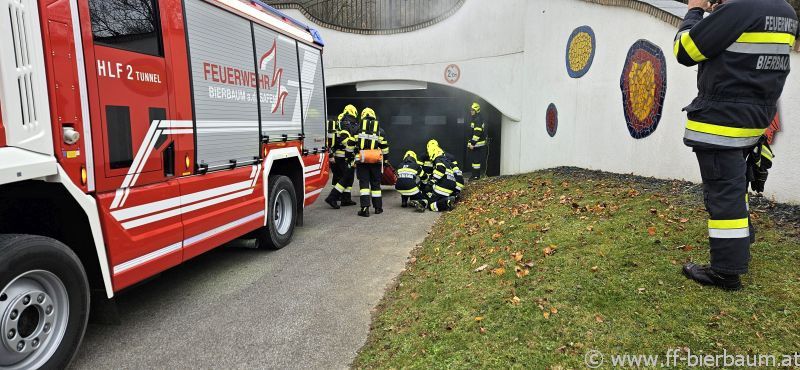 Feuerwehrleute kümmern sich um einen Notfall, wobei ein Feuerwehrwagen neben einem Tunneleingang geparkt ist. Sie befinden sich in der Nähe eines Rasenbereichs, und eine Person macht Fotos.