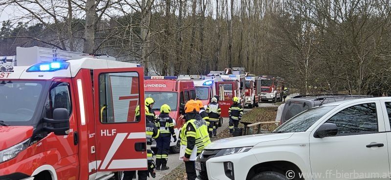 Eine Reihe von Rettungsfahrzeugen ist am Straßenrand geparkt, mit Feuerwehrleuten in gelben Helmen und Warnwesten. Bäume und ein weißes Auto sind im Hintergrund zu sehen.